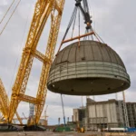 Une immense grue jaune à flèche treillis soulève un gigantesque dôme en acier au-dessus d’un vaste chantier industriel en béton, sous un ciel gris couvert.