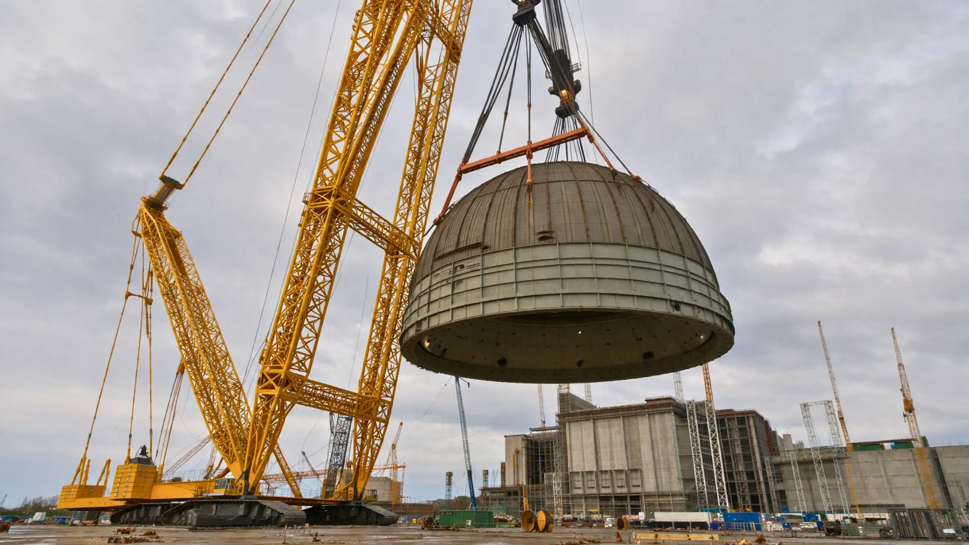 Une immense grue jaune à flèche treillis soulève un gigantesque dôme en acier au-dessus d’un vaste chantier industriel en béton, sous un ciel gris couvert.
