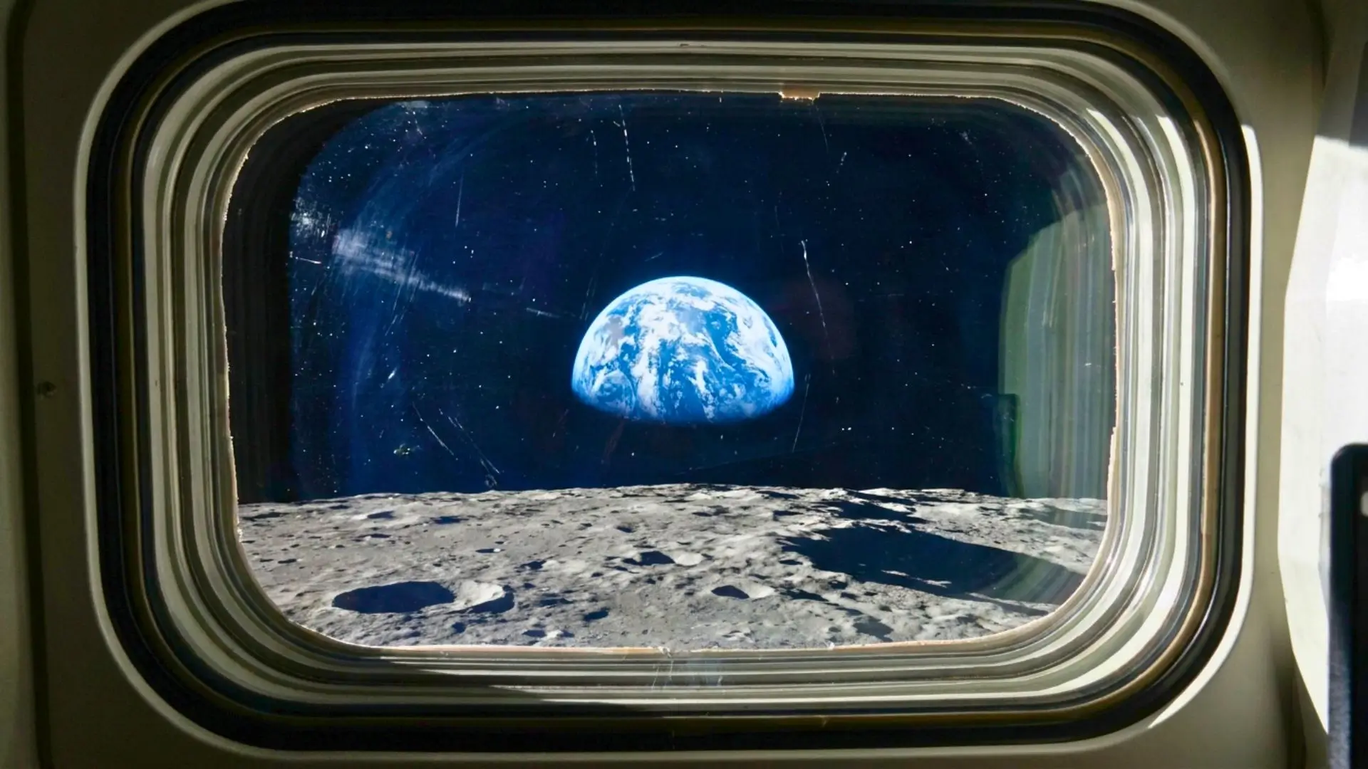 Vue à travers l'épais hublot d'un vaisseau spatial montrant la Terre bleue et blanche se levant au-dessus de la surface grise et cratérisée de la Lune.