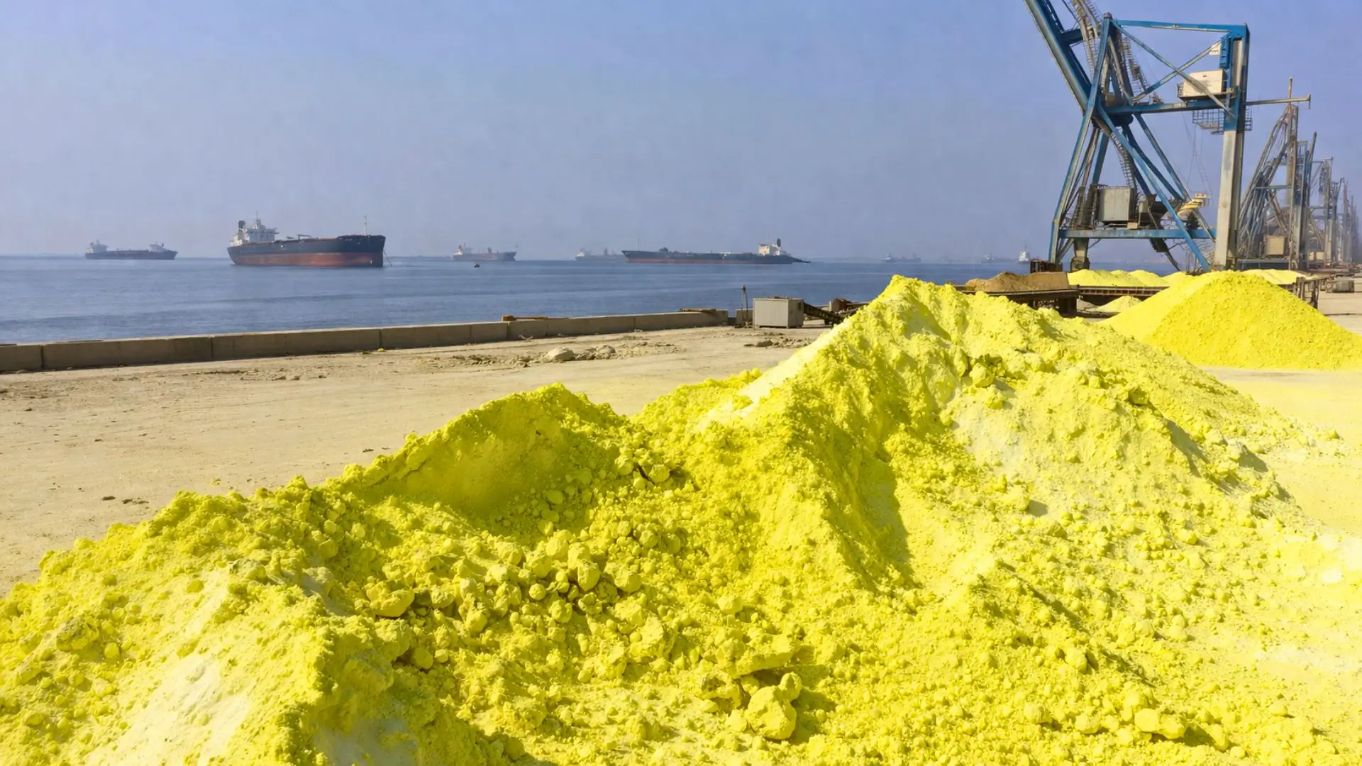 De vastes monticules de soufre jaune vif s’élèvent sur un quai industriel poussiéreux, face à des pétroliers immobiles sur une mer calme dans le détroit d’Ormuz.