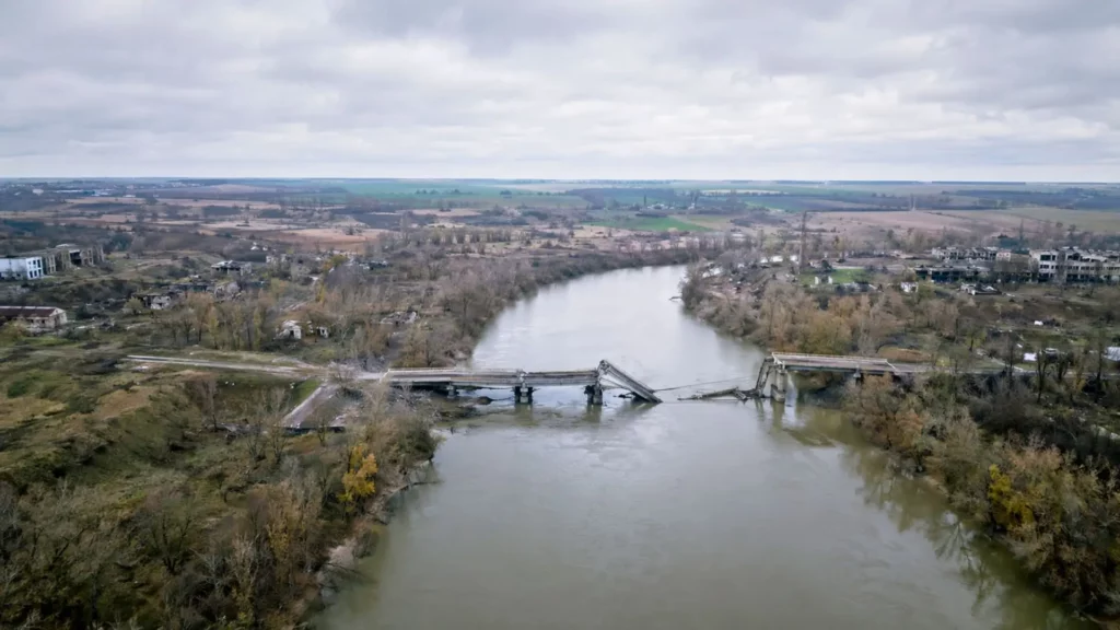 Vue aérienne d’un pont détruit traversant une large rivière boueuse, dans un paysage gris et désertique d’Europe de l’Est.