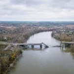 Vue aérienne d’un pont détruit traversant une large rivière boueuse, dans un paysage gris et désertique d’Europe de l’Est.