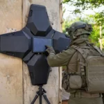Un opérateur militaire en tenue tactique manipule un radar noir en forme d’étoile fixé contre un mur en béton, en extérieur, sous la lumière du jour.