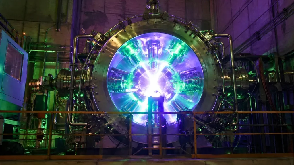 Une immense chambre à vide en acier émettant une lumière intense verte et violette, avec un cœur de plasma brillant au centre d’une installation industrielle.