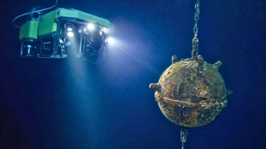 Un robot sous-marin télécommandé éclaire une mine navale rouillée suspendue dans les profondeurs obscures de l’océan.