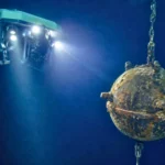 Un robot sous-marin télécommandé éclaire une mine navale rouillée suspendue dans les profondeurs obscures de l’océan.