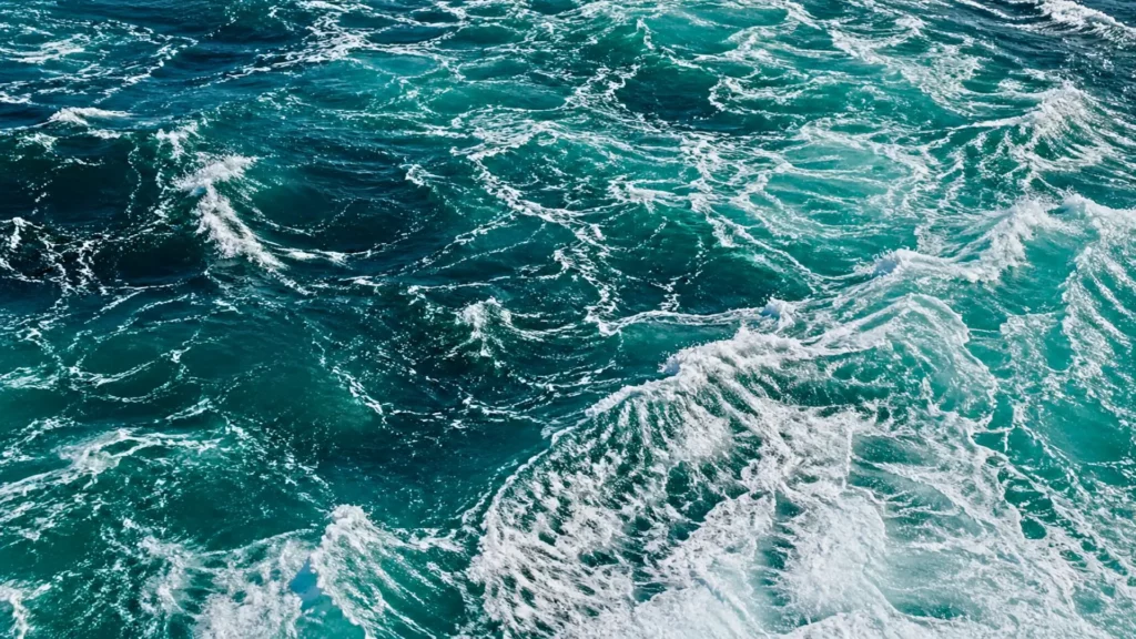 Vue aérienne d’une surface océanique agitée, avec de l’eau bleu-vert profonde et des motifs d’écume blanche tourbillonnants.