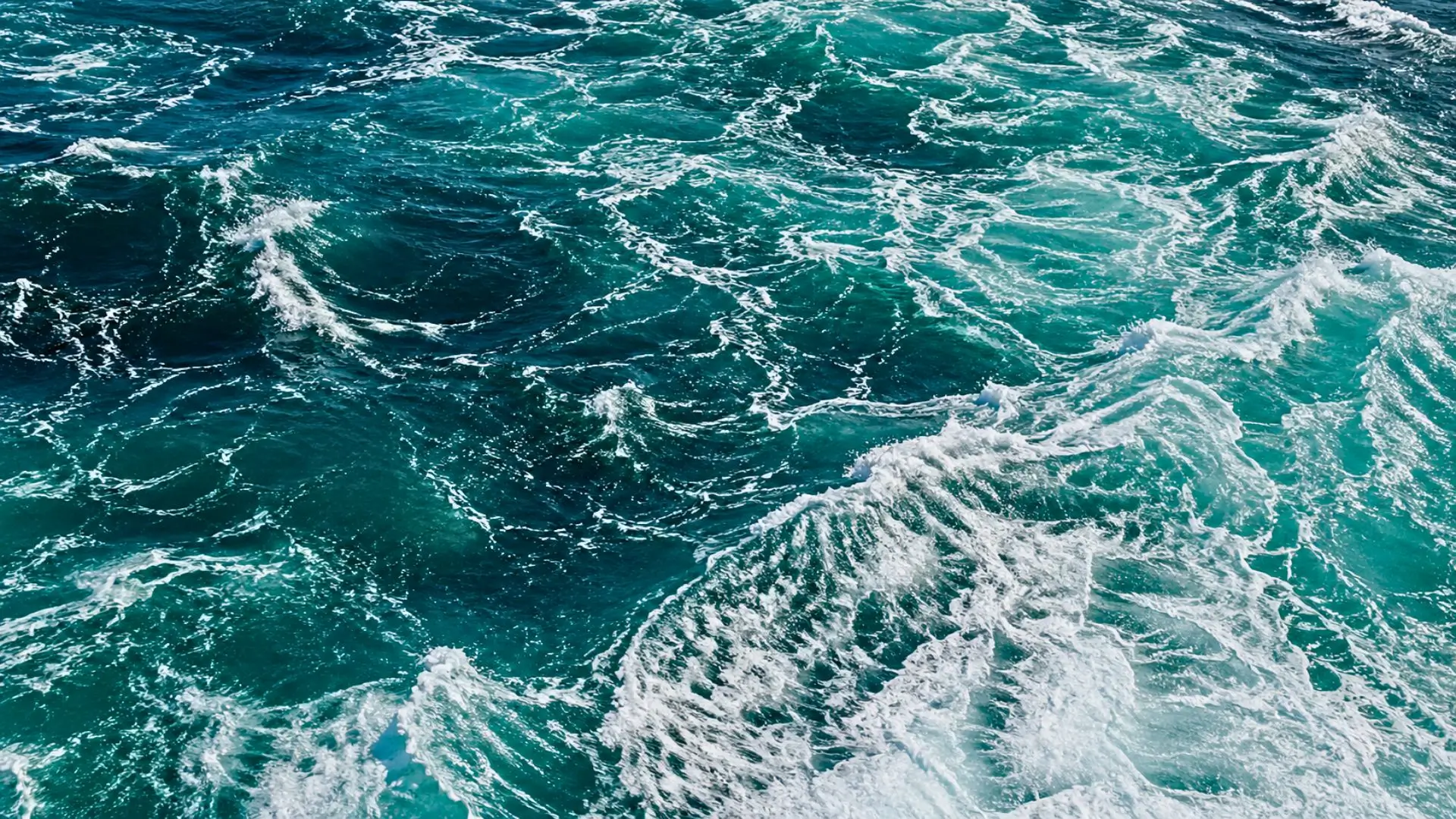 Vue aérienne d’une surface océanique agitée, avec de l’eau bleu-vert profonde et des motifs d’écume blanche tourbillonnants.