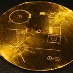 Gros plan photoréaliste du disque d’or de Voyager, posé sur une surface sombre, avec ses gravures scientifiques finement visibles et un reflet doré subtil.