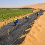 Barrière en fibre sombre installée entre dunes désertiques et champs cultivés.