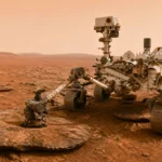 Rover Curiosity immobile sur la surface de Mars dans le cratère Gale, entouré d’un paysage rocheux et poussiéreux sous un ciel orangé