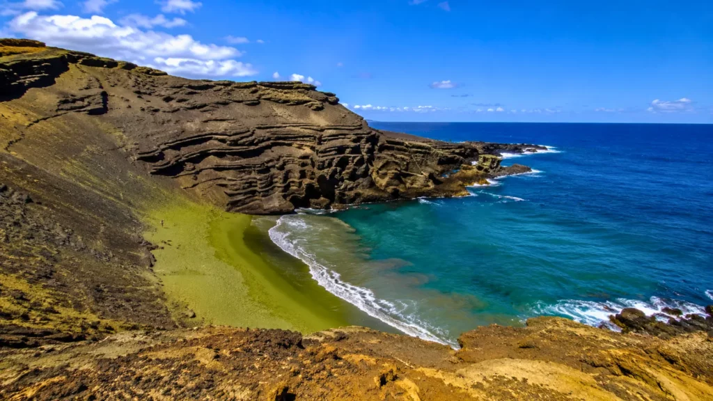 Randonneurs descendant vers la plage de sable vert Papakōlea à Hawaï, bordée de falaises volcaniques et d’une eau turquoise.