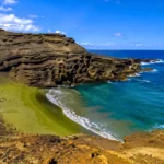 Randonneurs descendant vers la plage de sable vert Papakōlea à Hawaï, bordée de falaises volcaniques et d’une eau turquoise.