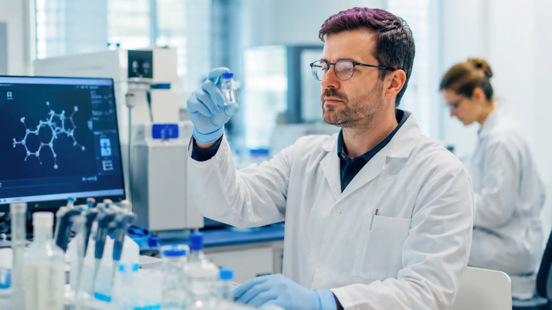 Chercheur en blouse blanche observant un flacon dans un laboratoire de recherche anticancer.