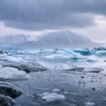 Pluie froide tombant sur une étendue glacée de l’Arctique avec des blocs de glace, de l’eau sombre et des montagnes sous un ciel couvert.