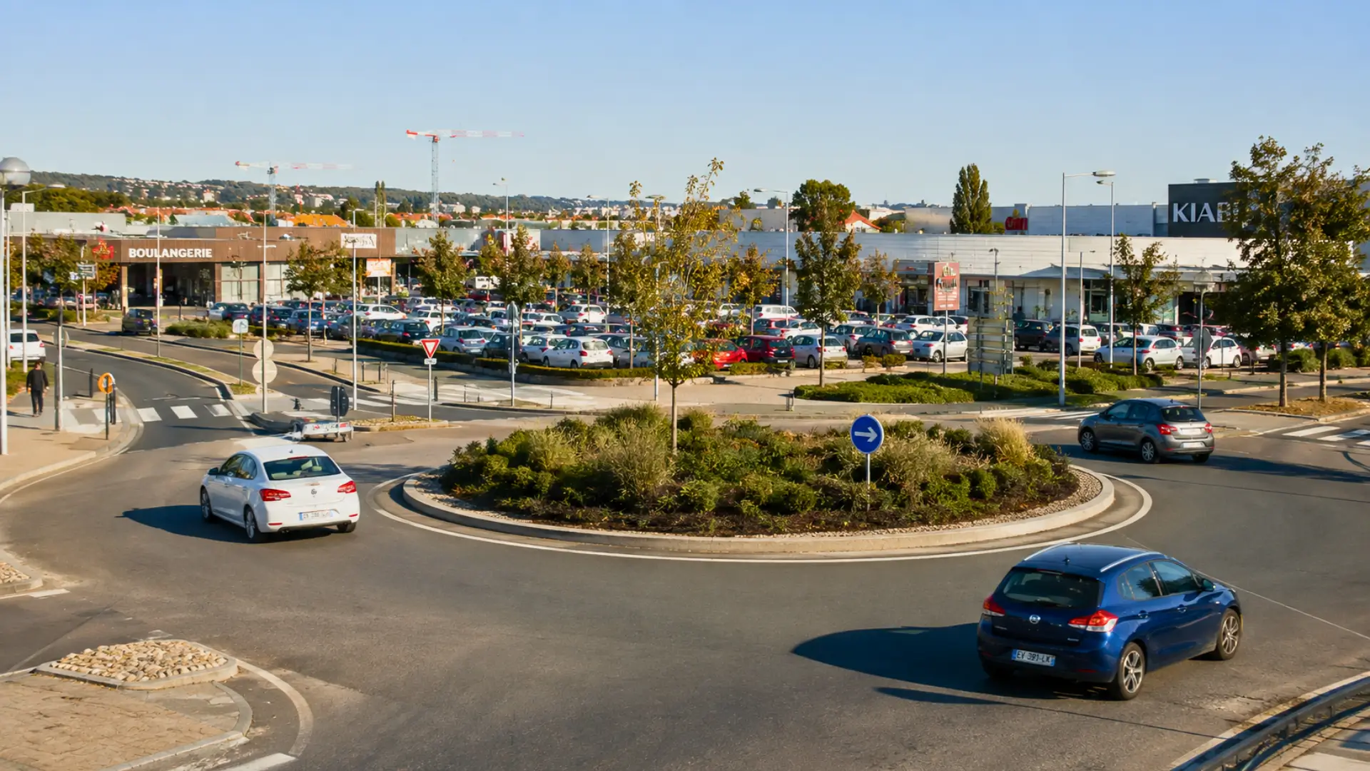 Rond-point paysager dans une zone commerciale française avec plusieurs voitures en circulation.