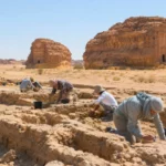 Des archéologues fouillent des vestiges enfouis dans le désert d’Hégra, avec des tombeaux nabatéens sculptés dans la roche en arrière-plan.