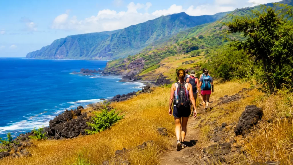 Randonneurs sur un sentier côtier facile à La Réunion, entre herbes dorées, roches volcaniques noires, montagnes verdoyantes et océan Indien.