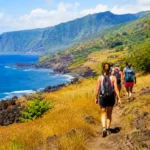 Randonneurs sur un sentier côtier facile à La Réunion, entre herbes dorées, roches volcaniques noires, montagnes verdoyantes et océan Indien.
