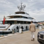 Un homme en tenue élégante marche vers un grand yacht de luxe amarré dans une marina, près d’une voiture haut de gamme.