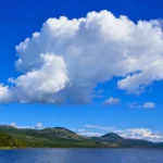 Grand cumulus blanc flottant dans un ciel bleu au-dessus d’un lac et de montagnes, illustrant le poids invisible des nuages.