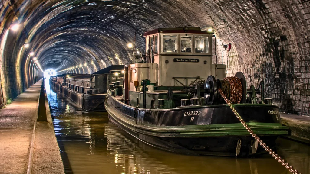 Un toueur électrique ancien remorque des péniches dans le tunnel de Riqueval, sous une voûte de pierre éclairée par des lampes.