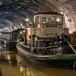 Un toueur électrique ancien remorque des péniches dans le tunnel de Riqueval, sous une voûte de pierre éclairée par des lampes.