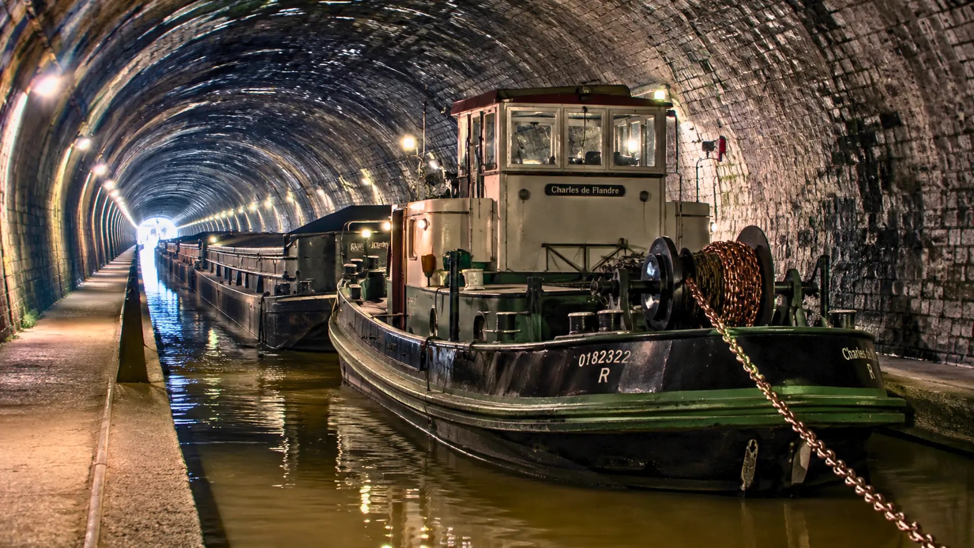 Un toueur électrique ancien remorque des péniches dans le tunnel de Riqueval, sous une voûte de pierre éclairée par des lampes.