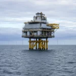 Plateforme industrielle offshore au large de la Manche, entourée par la mer, avec plusieurs éoliennes visibles en arrière-plan sous un ciel couvert.