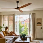 Ventilateur de plafond moderne dans un salon lumineux avec une femme assise près d’une fenêtre ouverte en été.