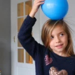 Une enfant tient un ballon bleu contre ses cheveux dans une pièce de la maison, illustrant naturellement un phénomène d’électricité statique.