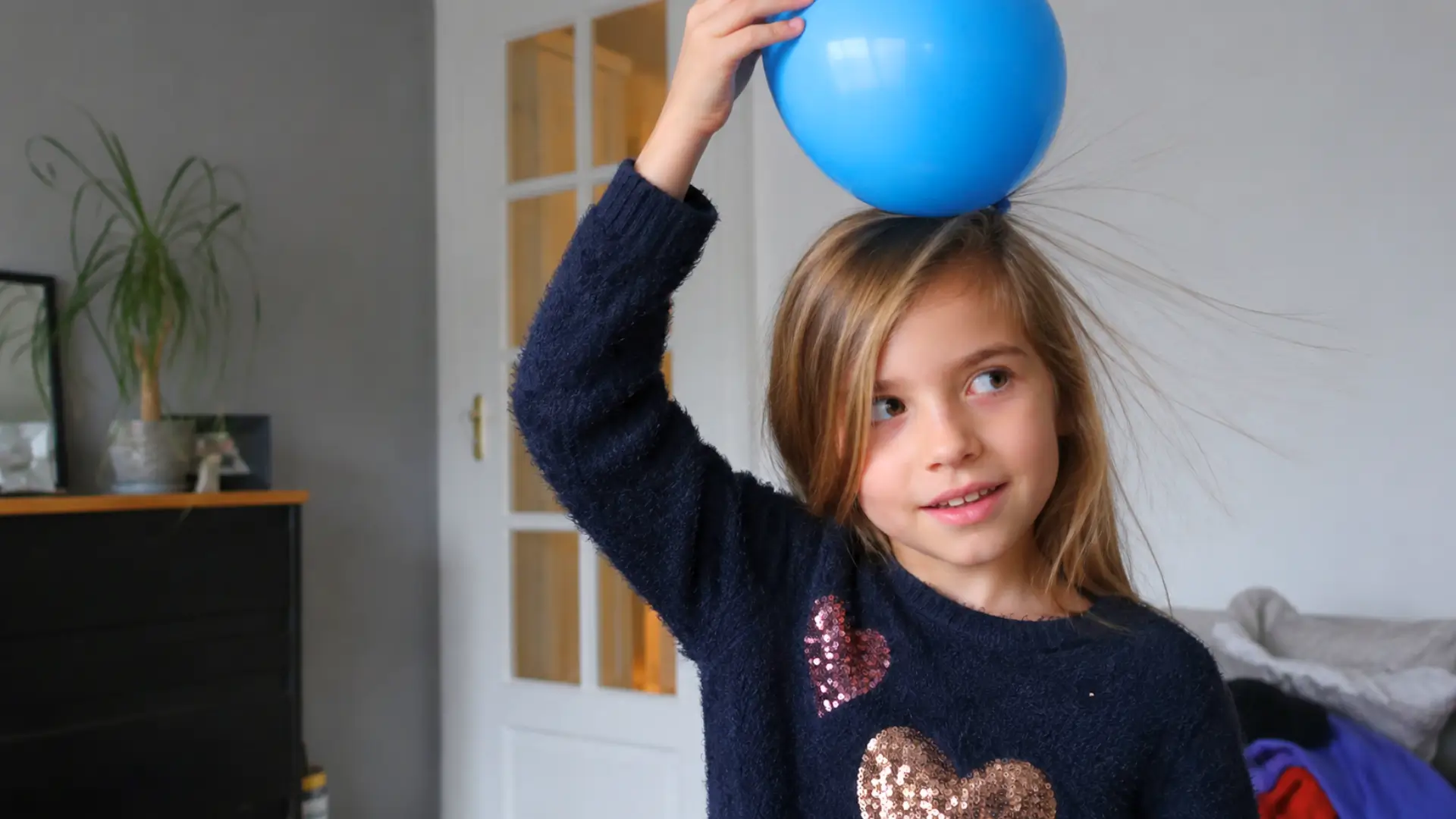 Une enfant tient un ballon bleu contre ses cheveux dans une pièce de la maison, illustrant naturellement un phénomène d’électricité statique.
