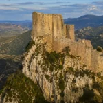 Une forteresse médiévale en ruines domine un éperon rocheux du Languedoc, au cœur d’un paysage montagneux.