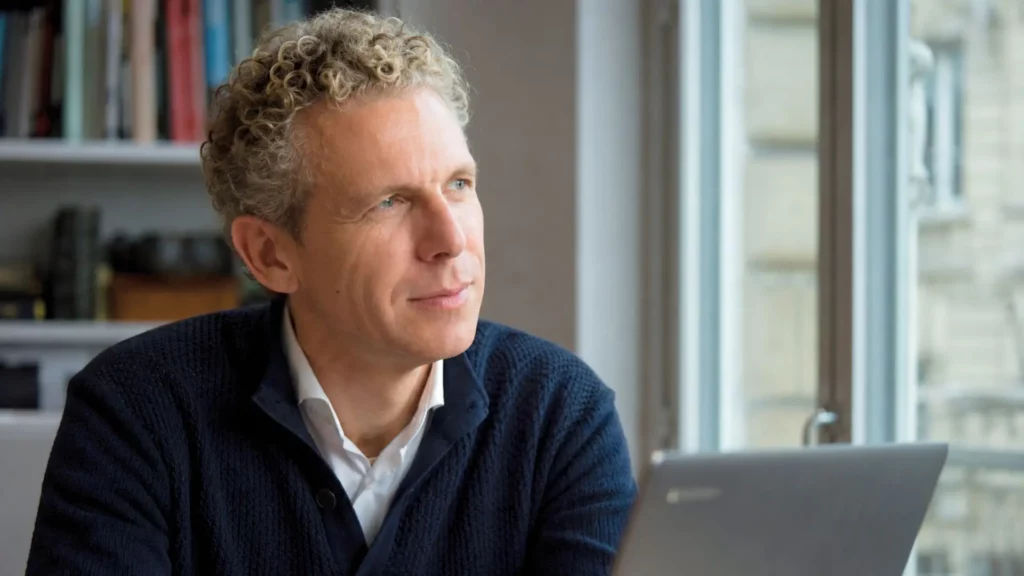 Gilles Babinet observe la lumière depuis son bureau, assis devant un ordinateur portable, dans un moment de réflexion sur l’intelligence artificielle.