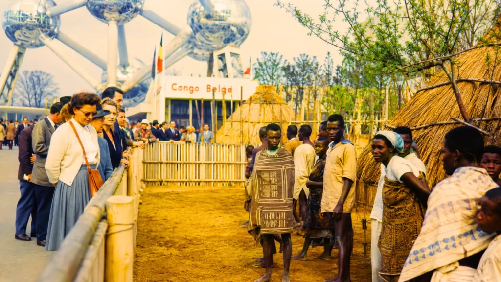 Reconstitution du village congolais de l’Expo 58 à Bruxelles, avec des visiteurs européens observant des Congolais derrière une clôture près de l’Atomium.