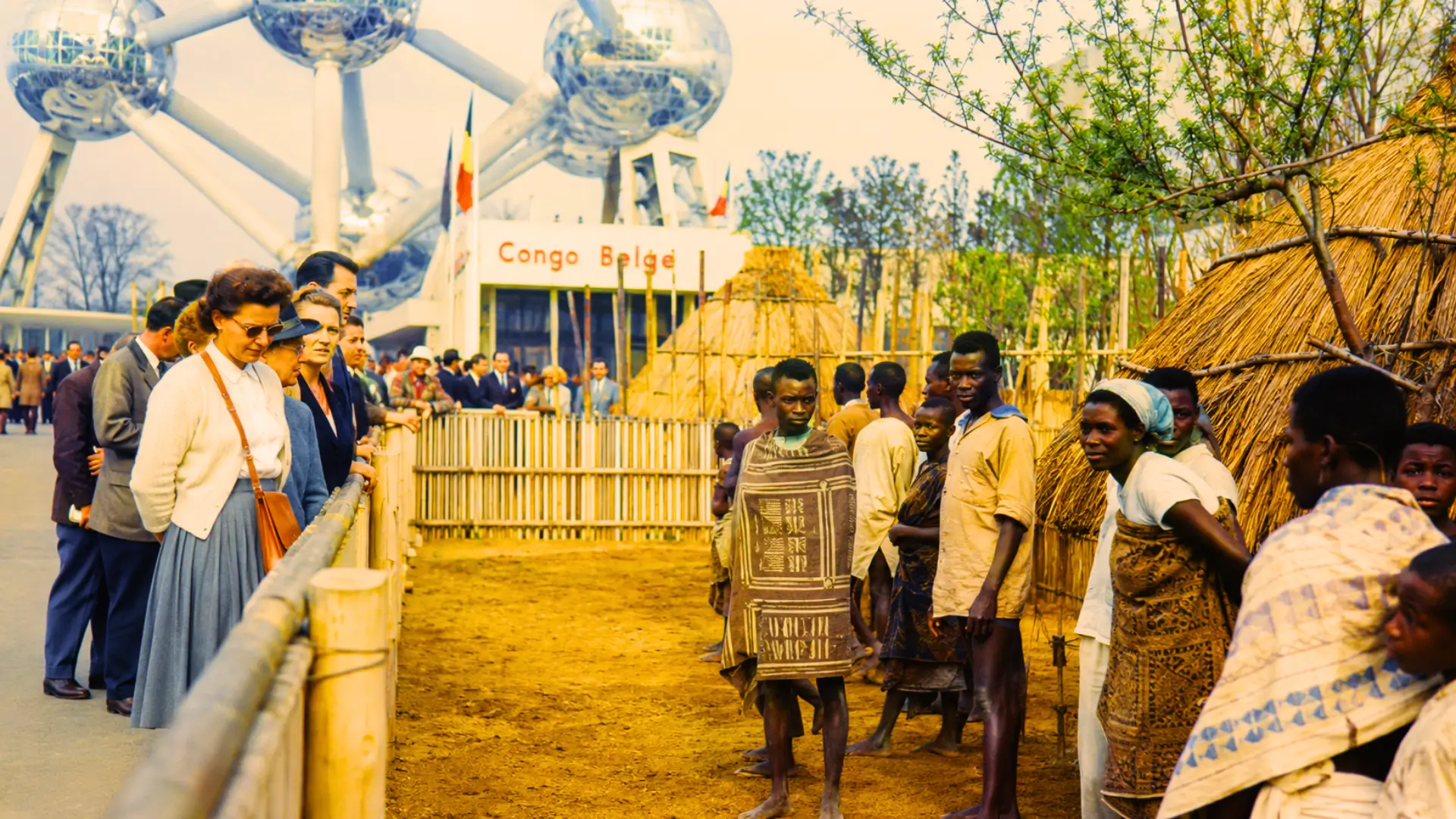Reconstitution du village congolais de l’Expo 58 à Bruxelles, avec des visiteurs européens observant des Congolais derrière une clôture près de l’Atomium.