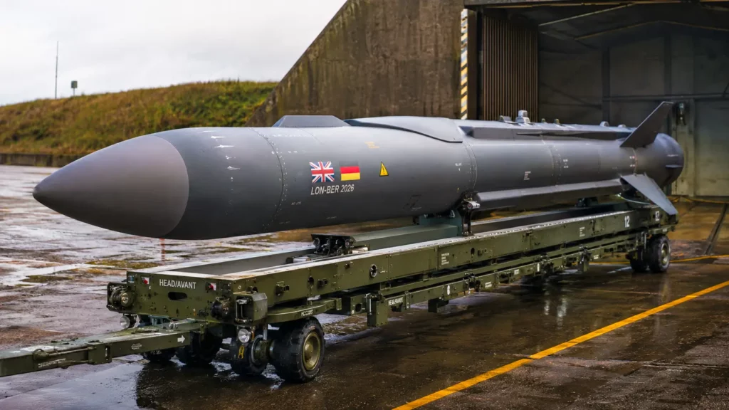 Un missile longue portée portant les drapeaux britannique et allemand repose sur un rail de transport devant un hangar militaire.