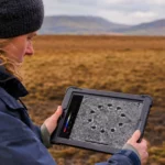 Une archéologue observe sur une tablette une carte géophysique révélant un cercle préhistorique enfoui sous la tourbe à Machrie Moor, en Écosse.