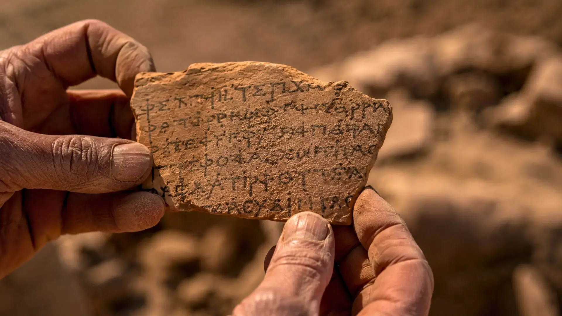 Un fragment de céramique inscrit est tenu entre des mains poussiéreuses sur un site de fouilles en Égypte.