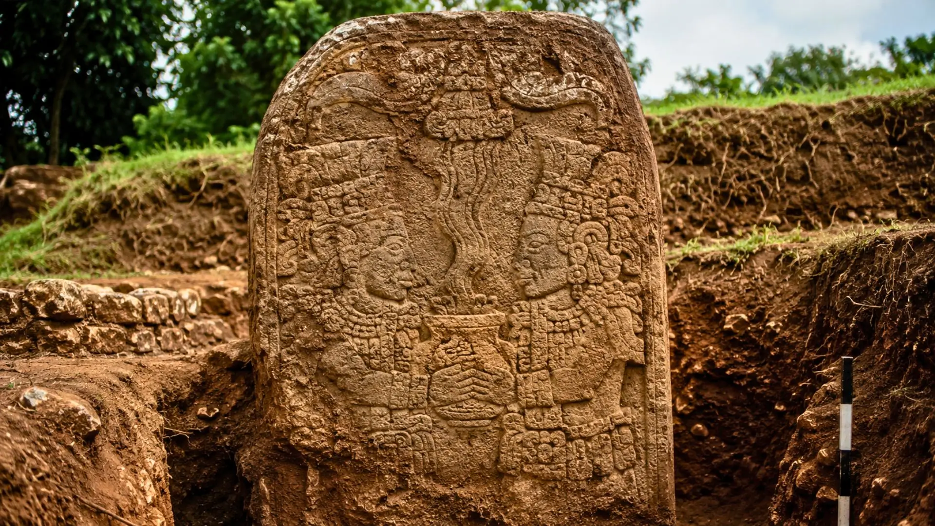 Une stèle mexicaine ancienne, sculptée de deux dignitaires face à face autour d’un récipient rituel, apparaît au centre d’une fouille archéologique.