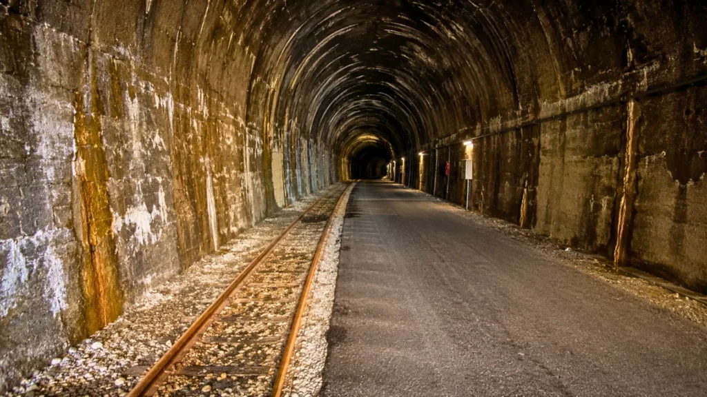 Les anciens rails du tunnel ferroviaire du Somport longent une chaussée asphaltée éclairée dans l’obscurité.