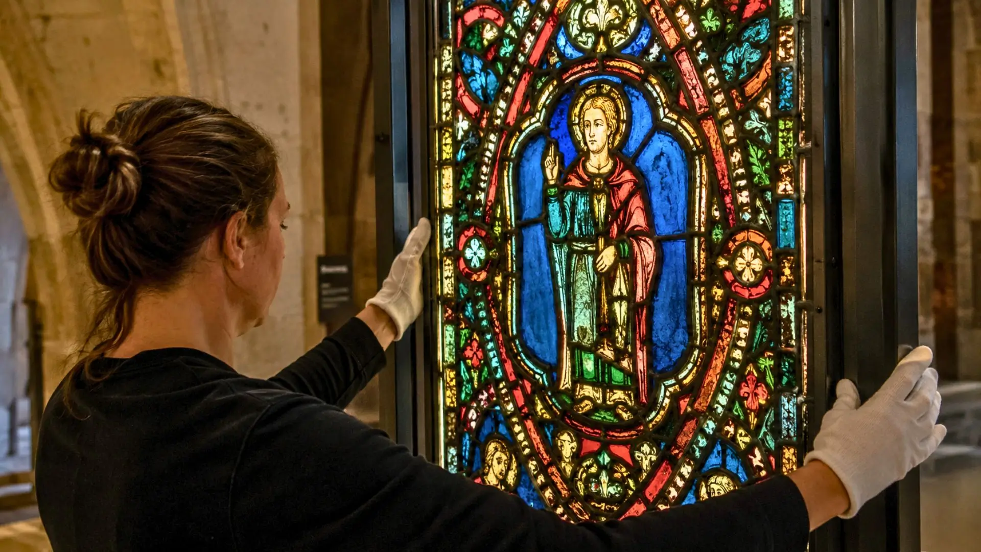 Une conservatrice gantée examine un vitrail médiéval aux couleurs éclatantes dans la Cité du Vitrail à Troyes.