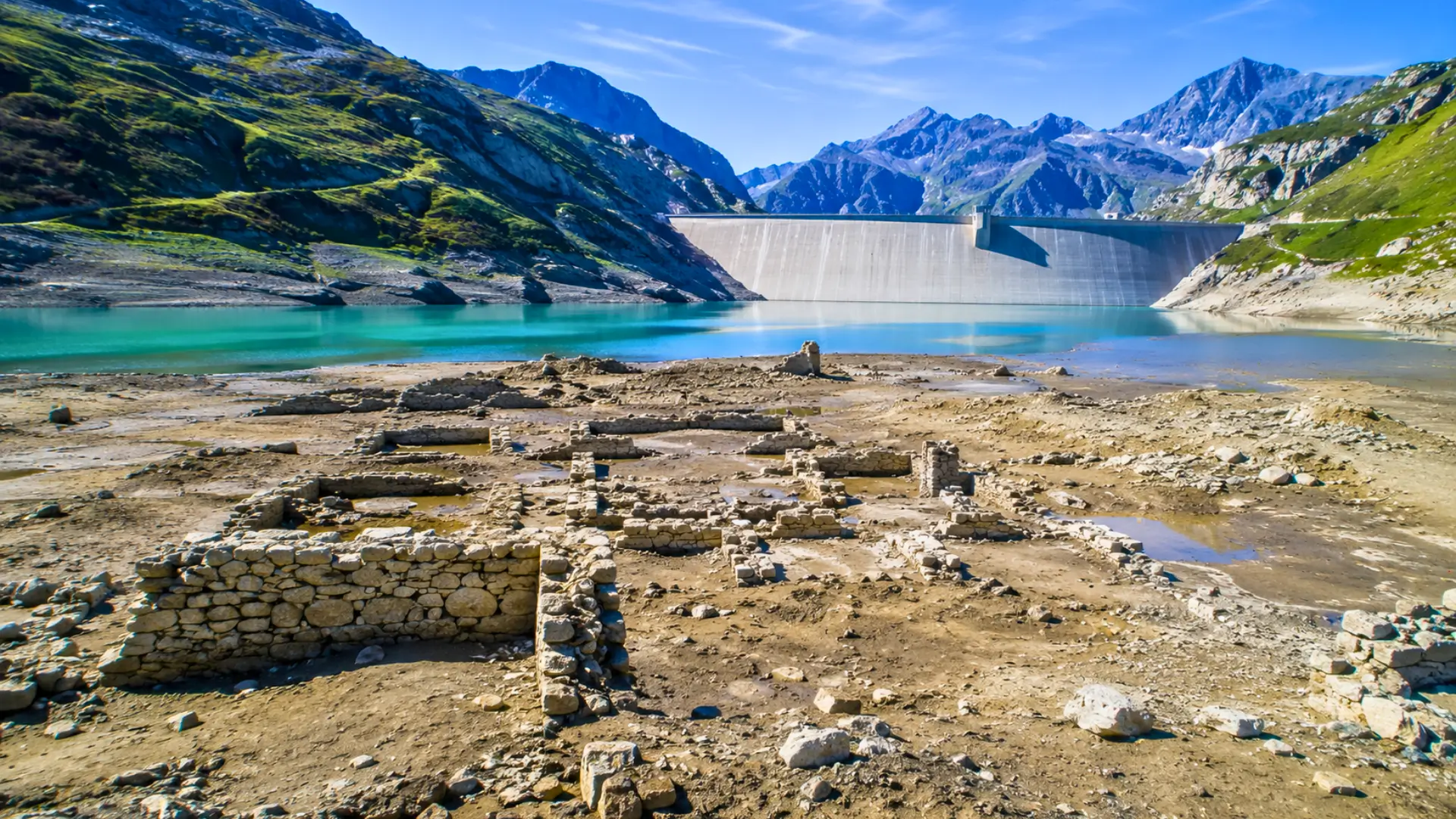 Vestiges du Vieux Tignes visibles sur les rives du lac du Chevril, avec le barrage et les montagnes alpines en arrière-plan.