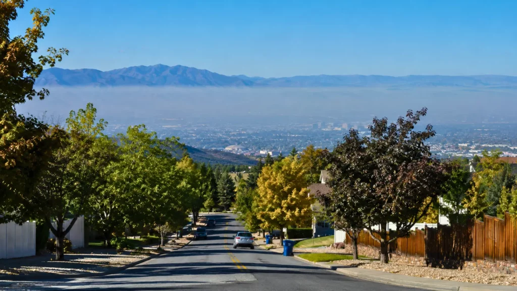 Rue résidentielle en pente bordée d’arbres, sous un ciel bleu clair, avec une fine couche de pollution atmosphérique au-dessus d’une vallée et des montagnes au loin après des incendies.