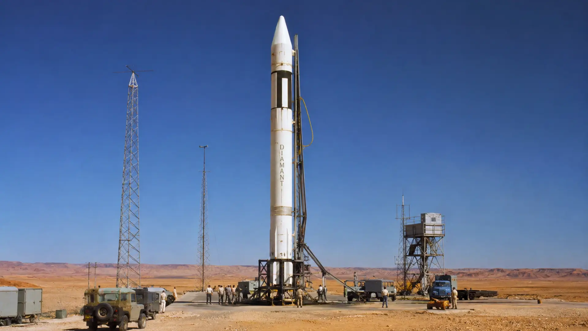 La fusée française Diamant-A dressée sur son pas de tir à Hammaguir, dans le Sahara algérien, entourée d’antennes, de véhicules techniques et de techniciens.