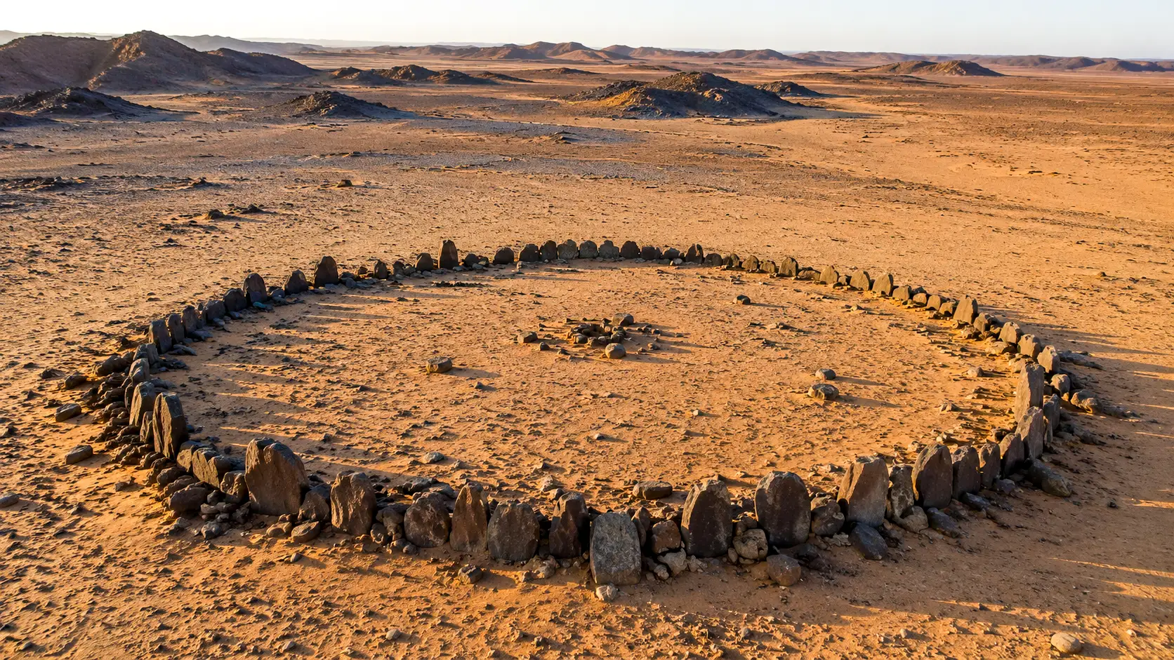 Un vaste cercle de pierres préhistorique se détache dans le désert d’Atbai, entre étendues arides et reliefs rocheux.
