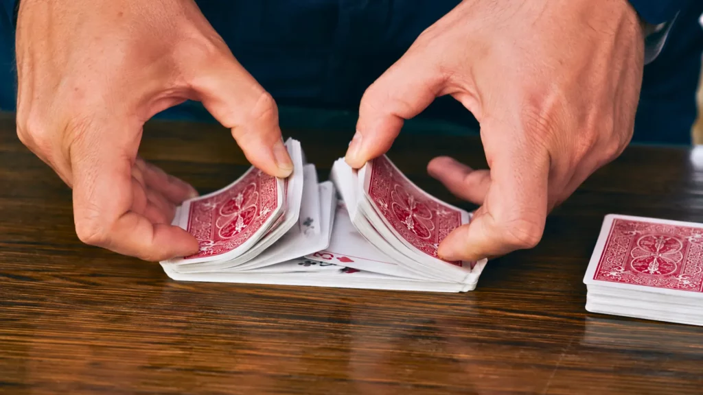 Des mains effectuent un riffle shuffle avec un jeu de 52 cartes sur une table en bois.