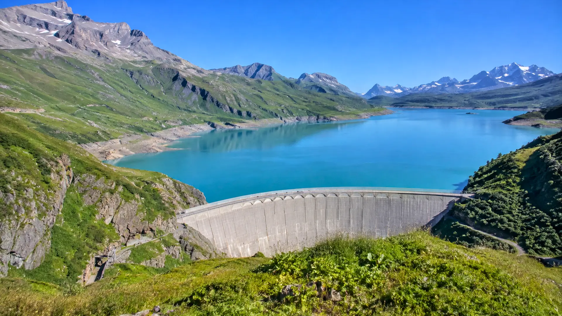 Vue panoramique du barrage de Roselend retenant un lac turquoise dans le massif du Beaufortain, entouré de montagnes alpines sous un ciel dégagé.