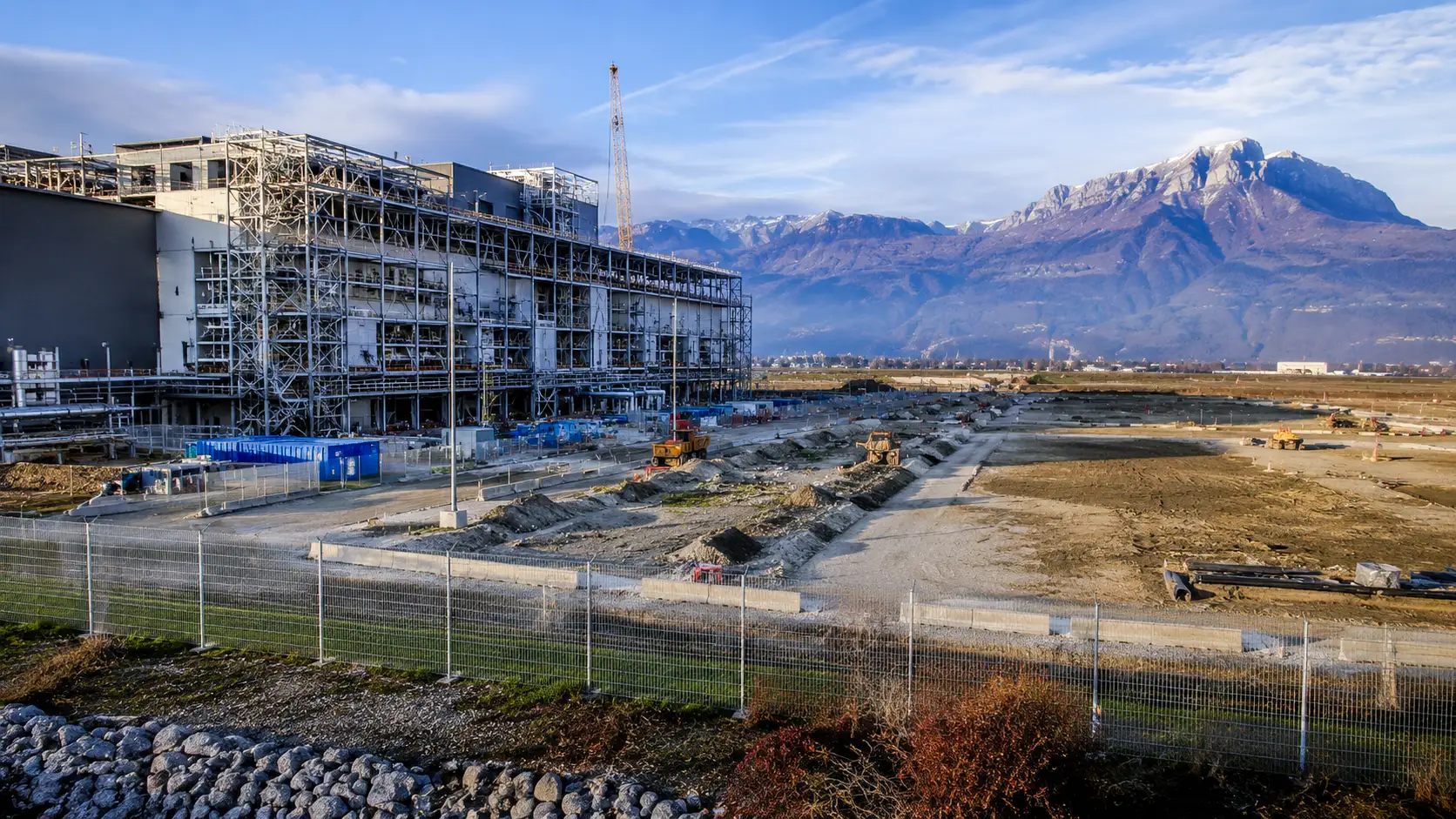 Chantier d’extension d’une usine de semi-conducteurs à Crolles, avec un bâtiment industriel en construction et une vaste zone encore inoccupée au pied des Alpes.
