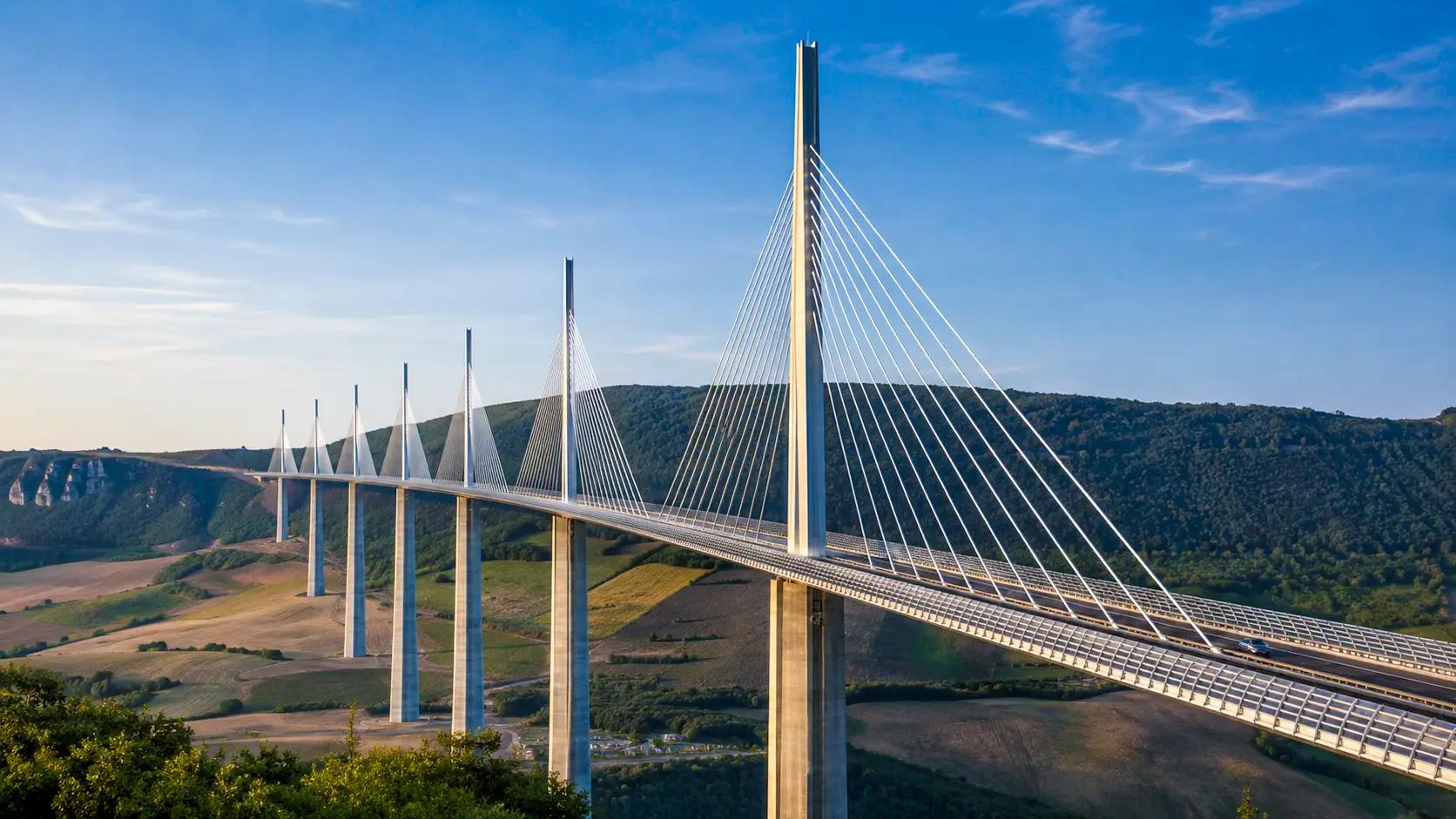 Le viaduc de Millau traverse la vallée du Tarn, ses immenses pylônes et haubans dominant le paysage sous une lumière dorée.